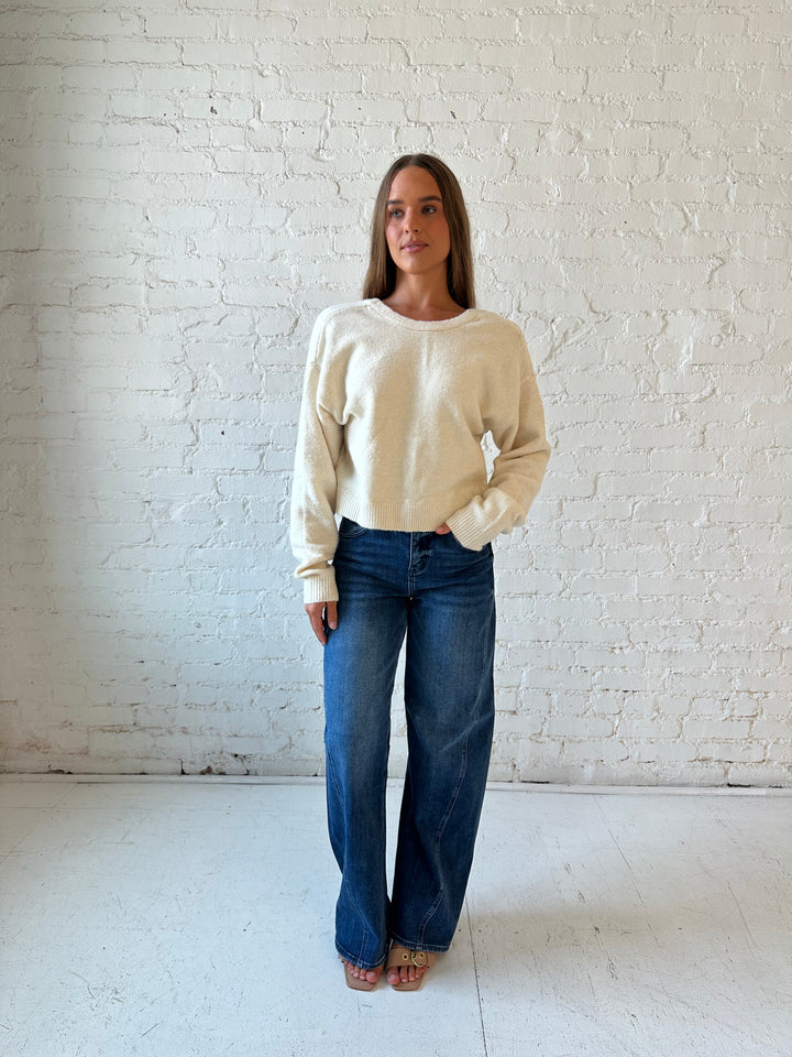 Woman wearing cream reversible bow sweater with dark wash barrel jean. 