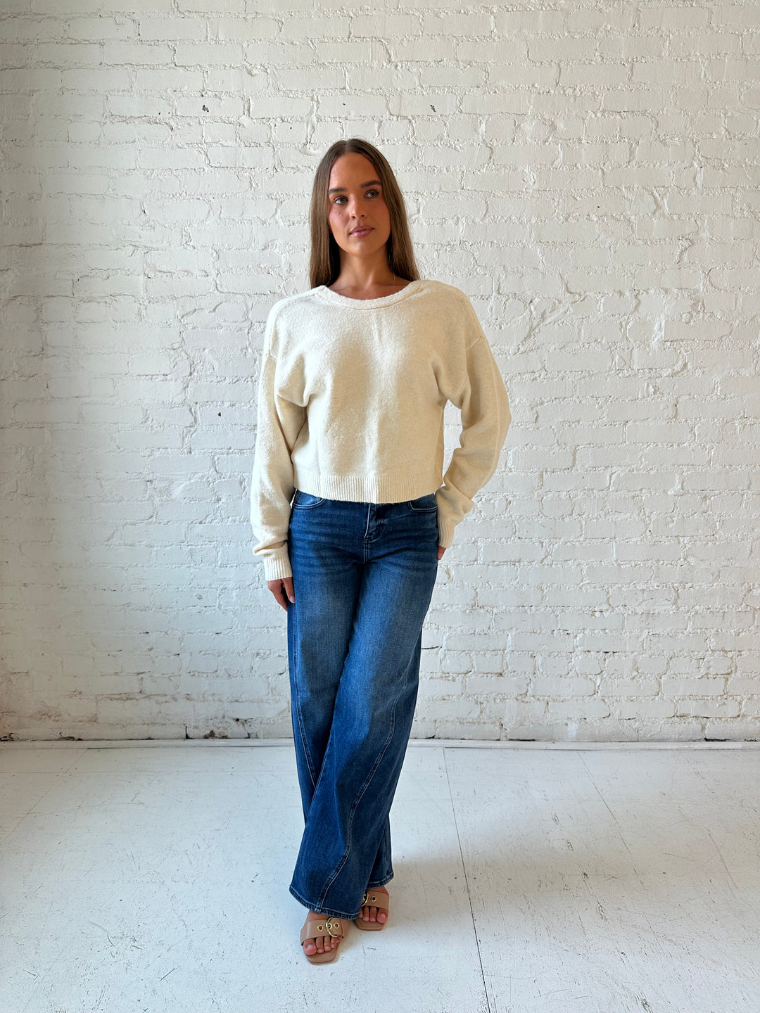 Woman wearing cream reversible bow sweater with dark wash barrel jean. 