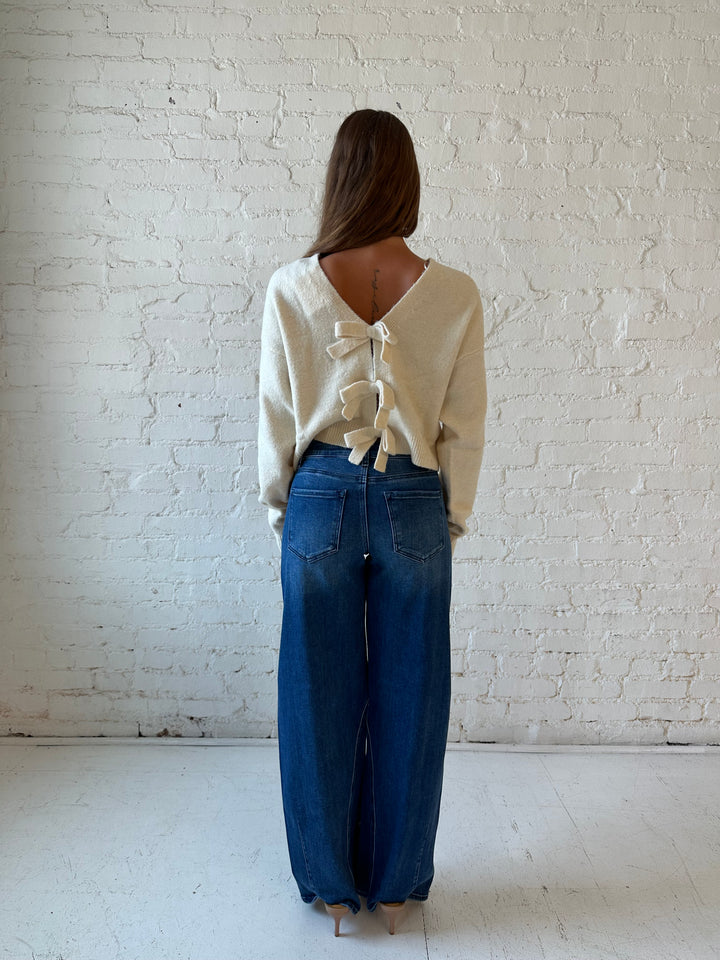 Woman wearing cream reversible bow sweater with dark wash barrel jean. 