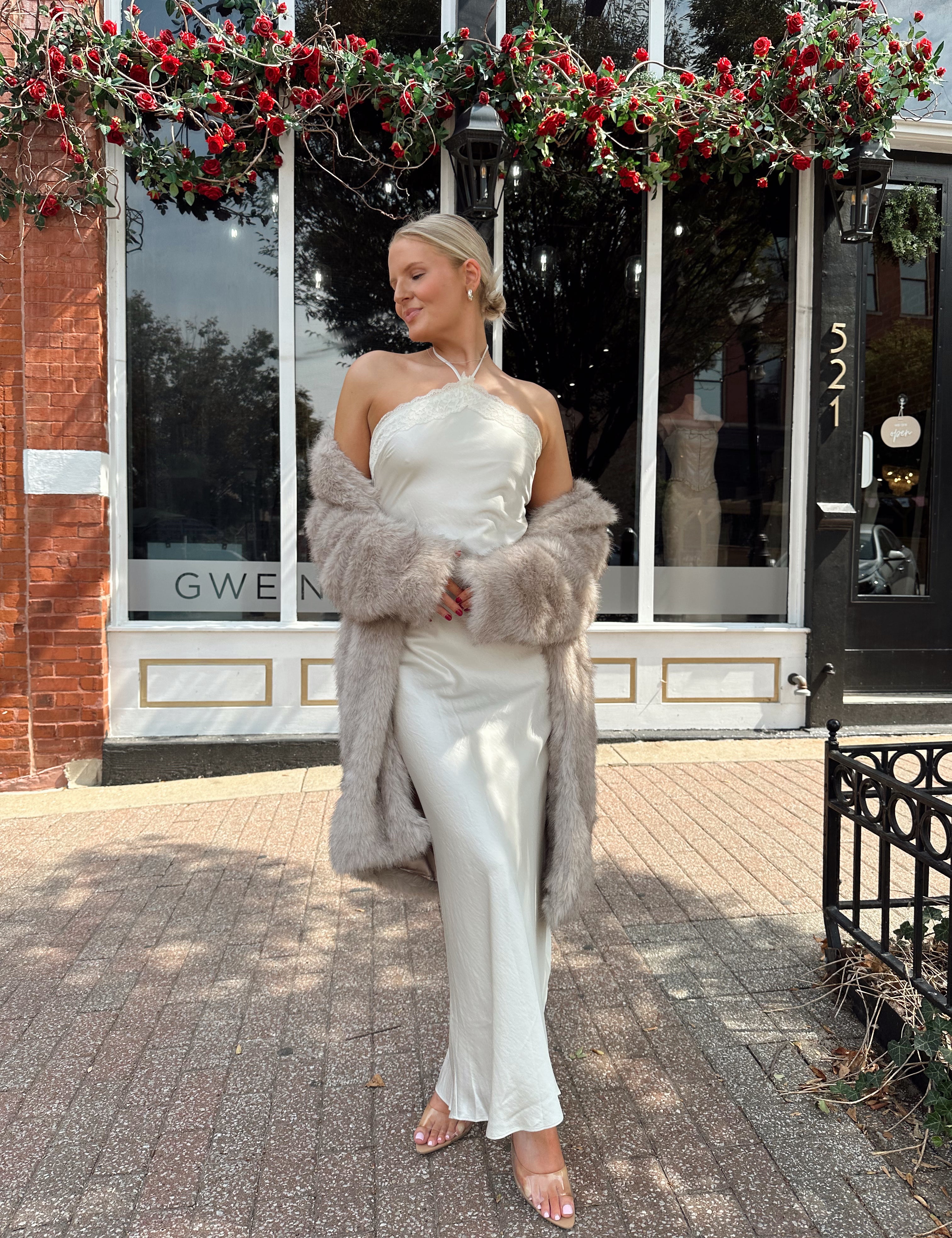 Woman wearing beige faux fur coat over white satin slip dress.