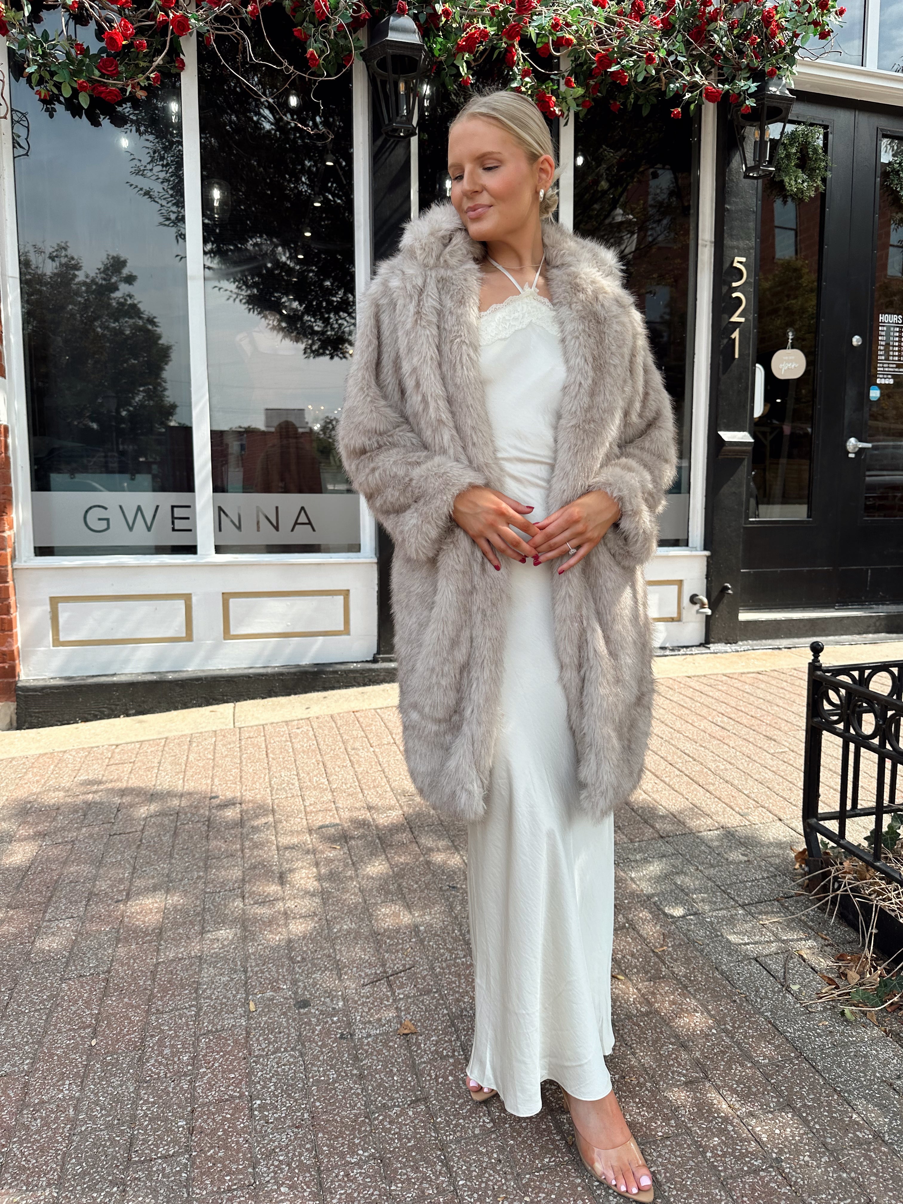 Woman wearing beige faux fur coat over white satin slip dress.
