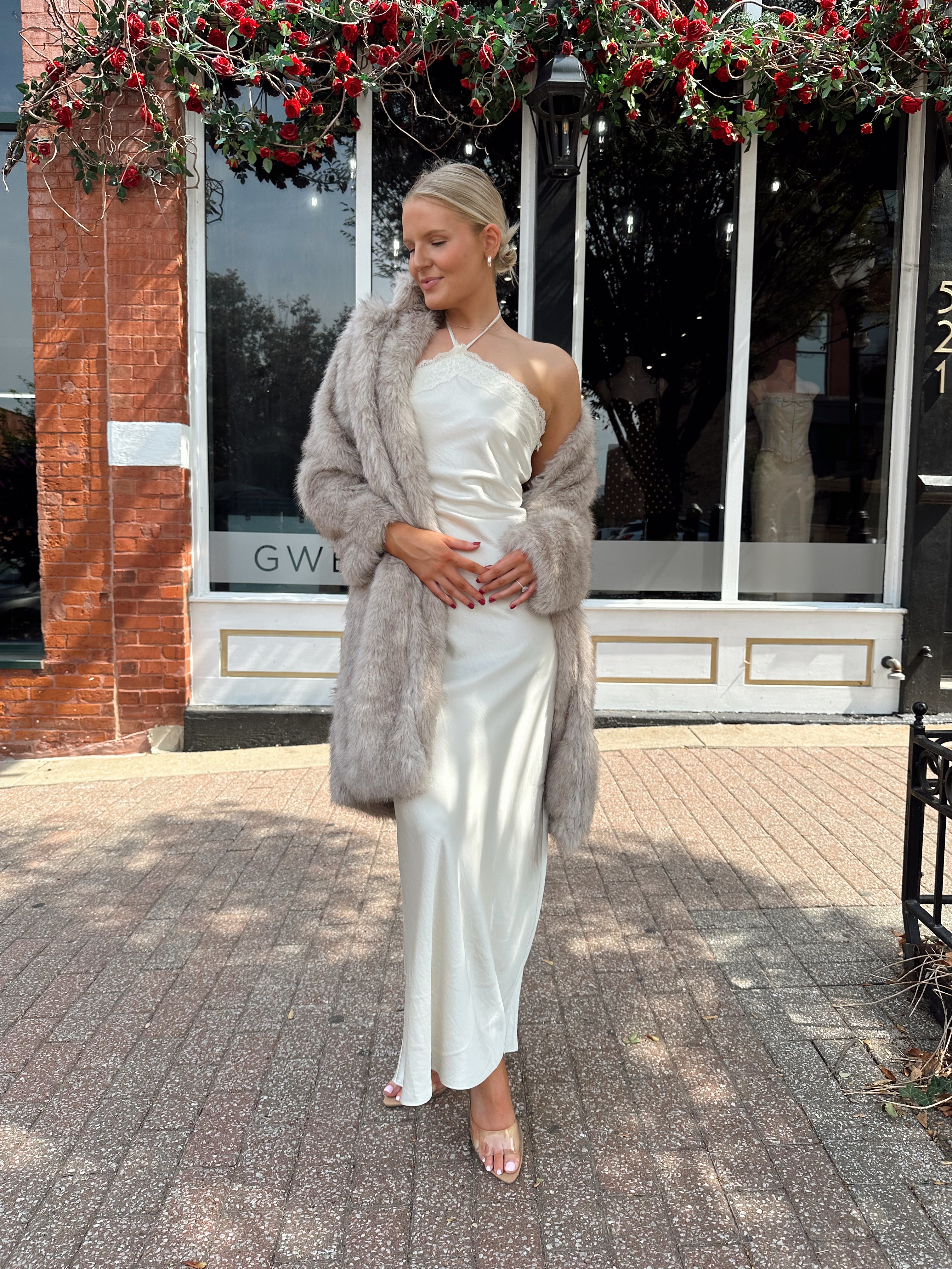 Woman wearing beige faux fur coat over white satin slip dress.