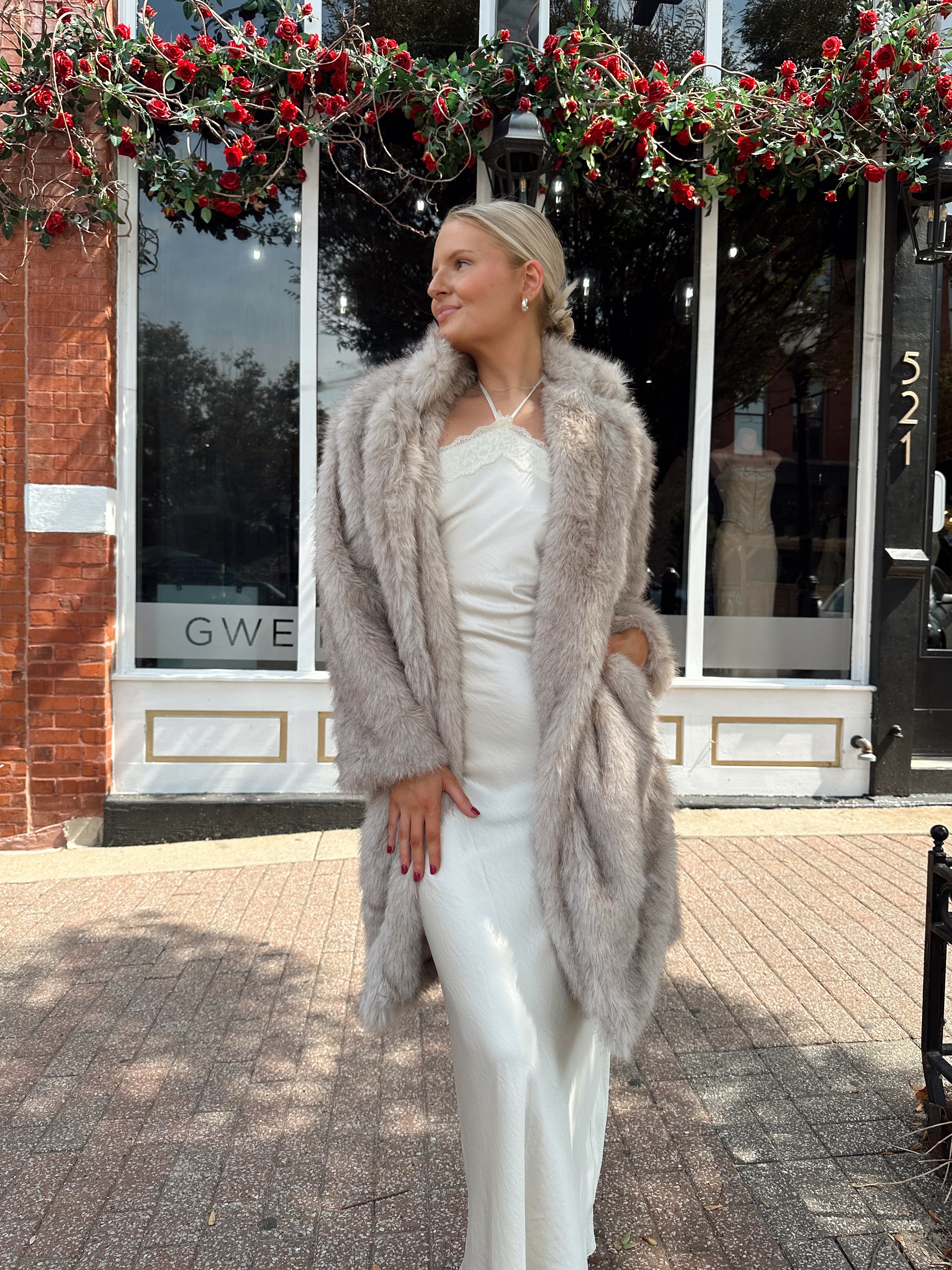 Woman wearing beige faux fur coat over white satin slip dress.