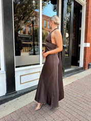 Woman wearing chocolate brown satin maxi dress with open back and long ties. 