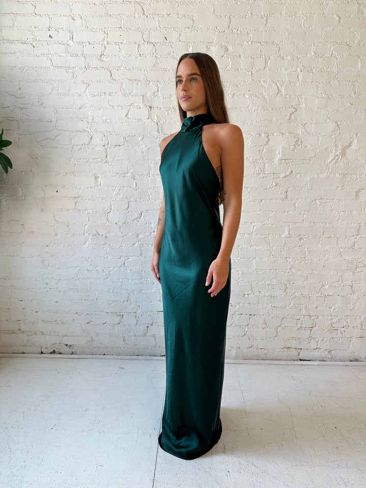 Woman wearing emerald green satin maxi dress with high self tie halter neckline. 