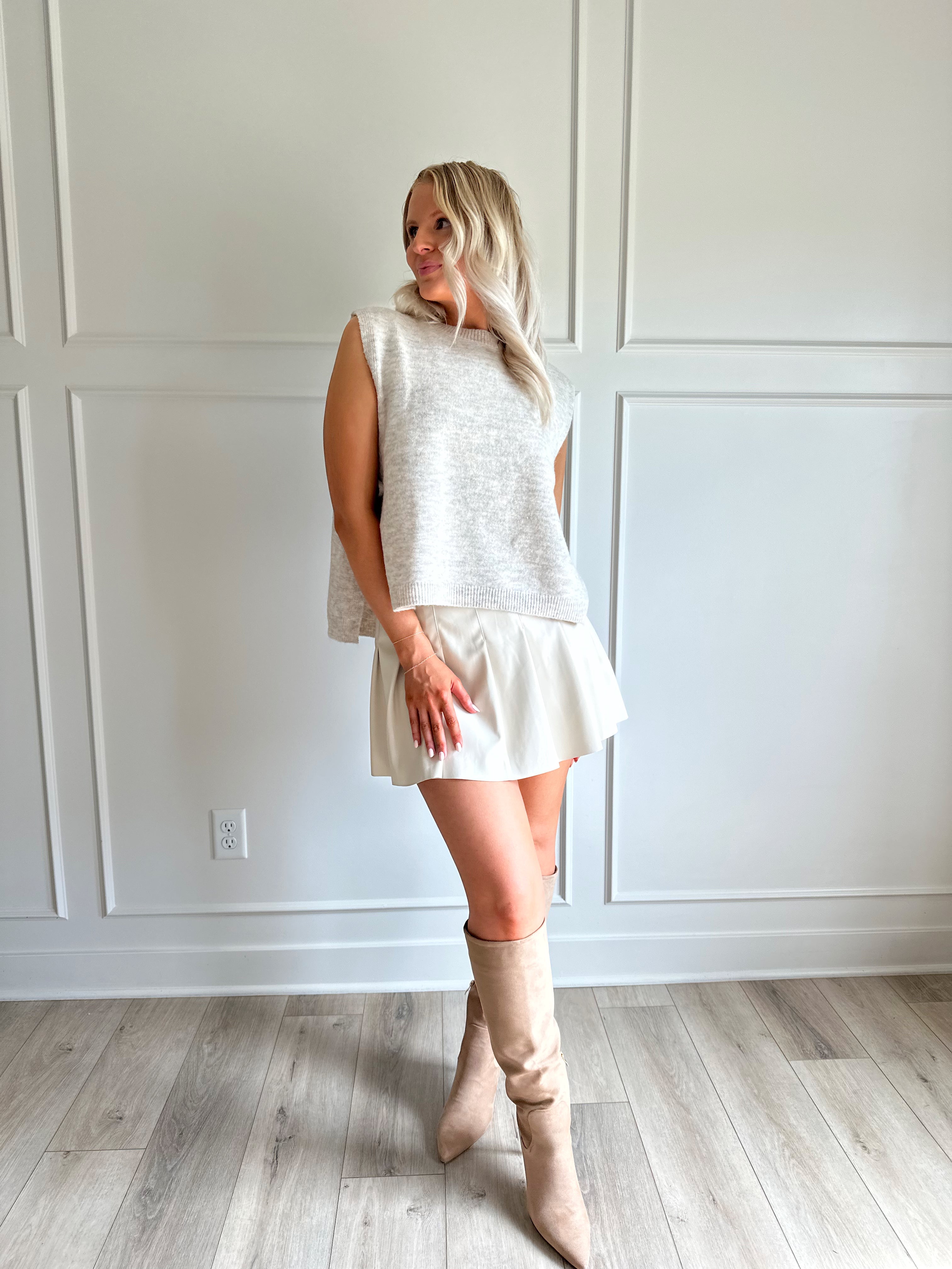 Woman wearing a cream vegan leather pleated mini skirt and oatmeal knit sleeveless top. 