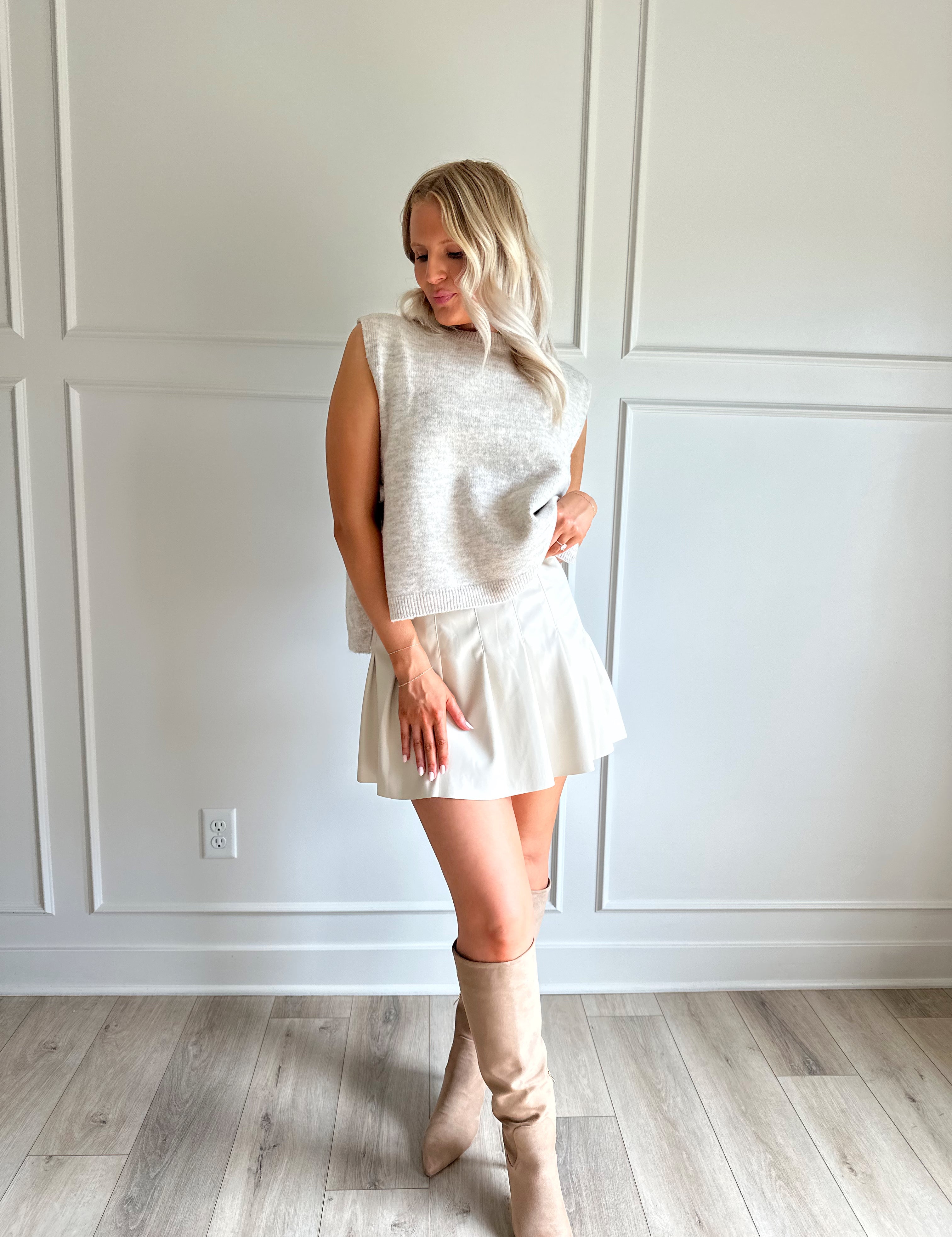 Woman wearing an oatmeal sleeveless knit top with side tie detail, styled with a pleated skirt and boots.
