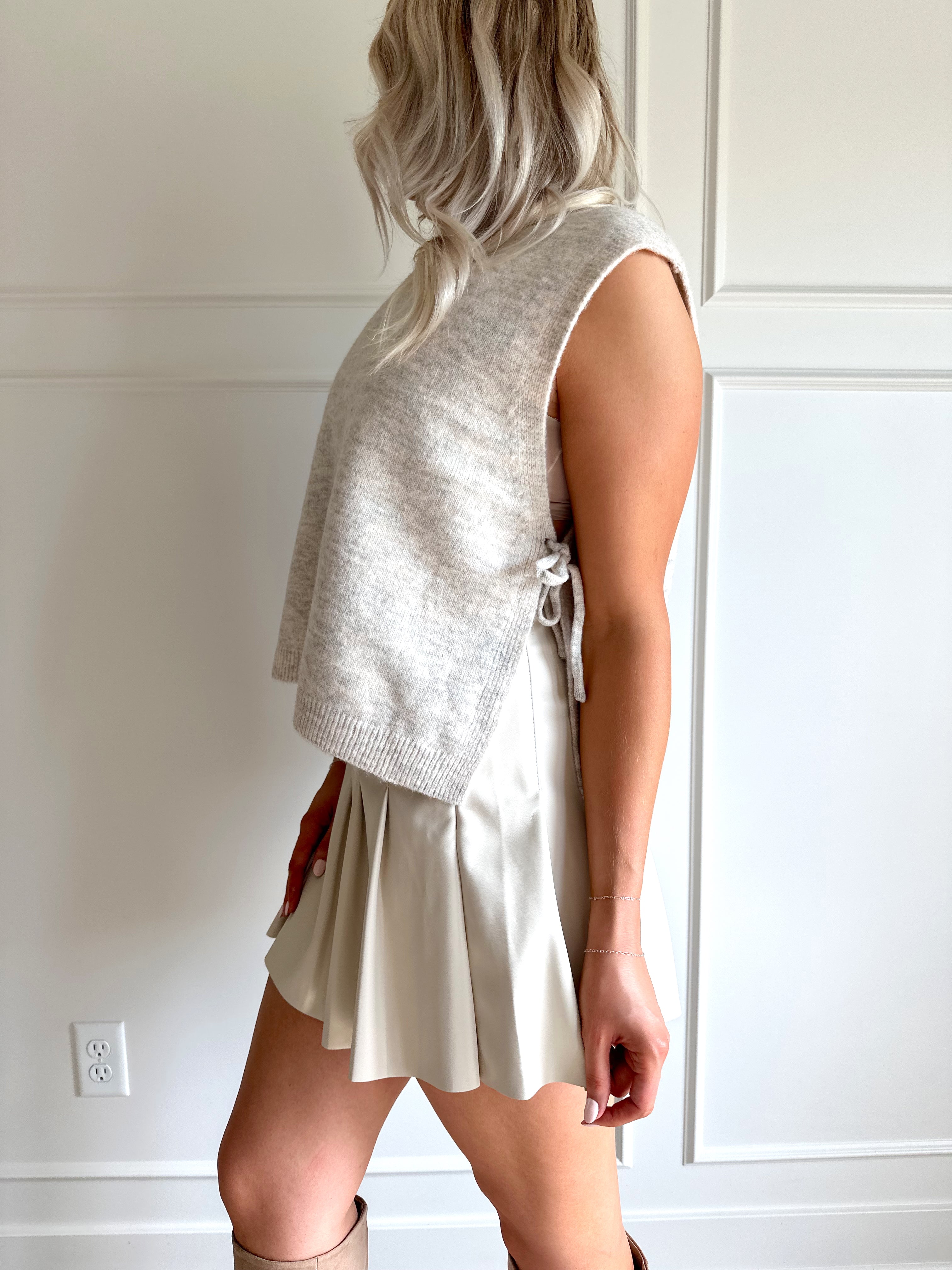 Woman wearing an oatmeal sleeveless knit top with side tie detail, styled with a pleated skirt and boots.