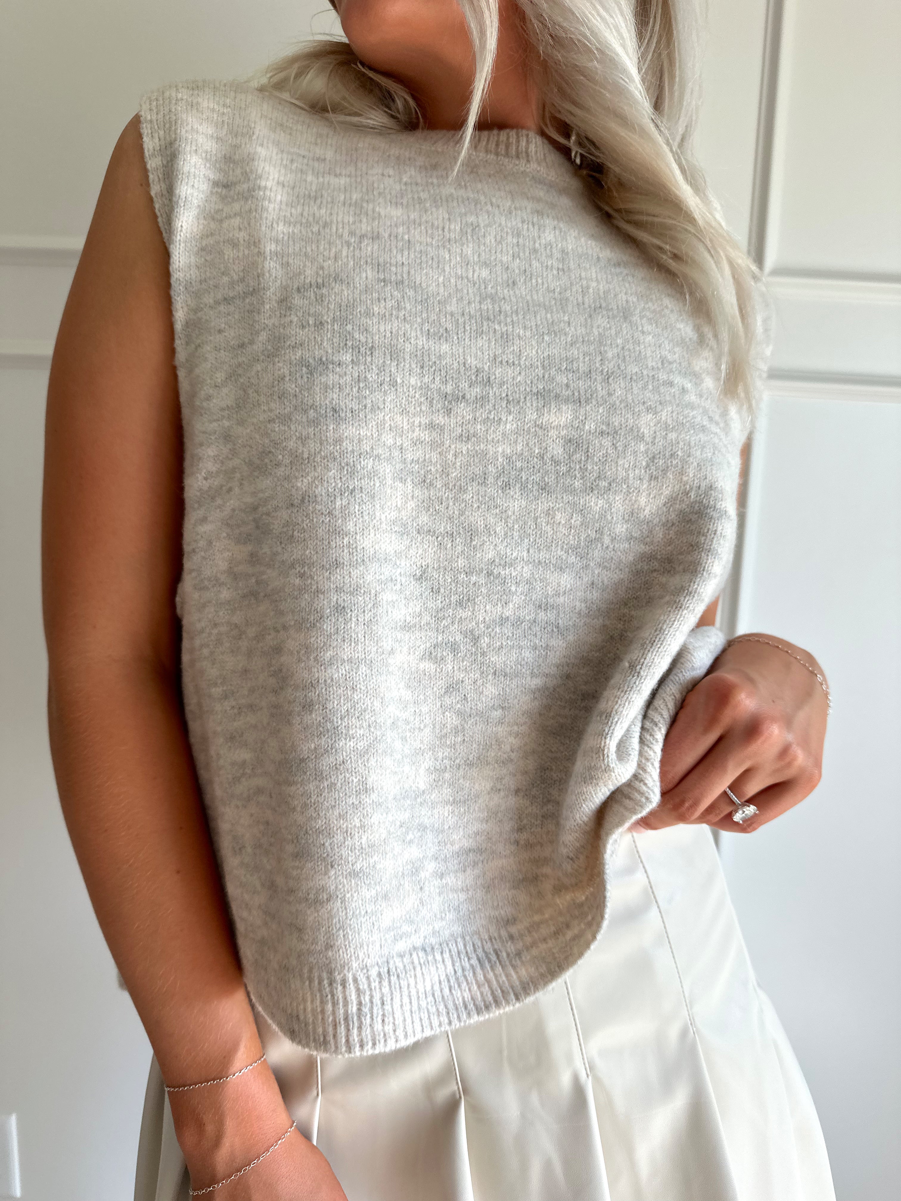Woman wearing an oatmeal sleeveless knit top with side tie detail, styled with a pleated skirt and boots.