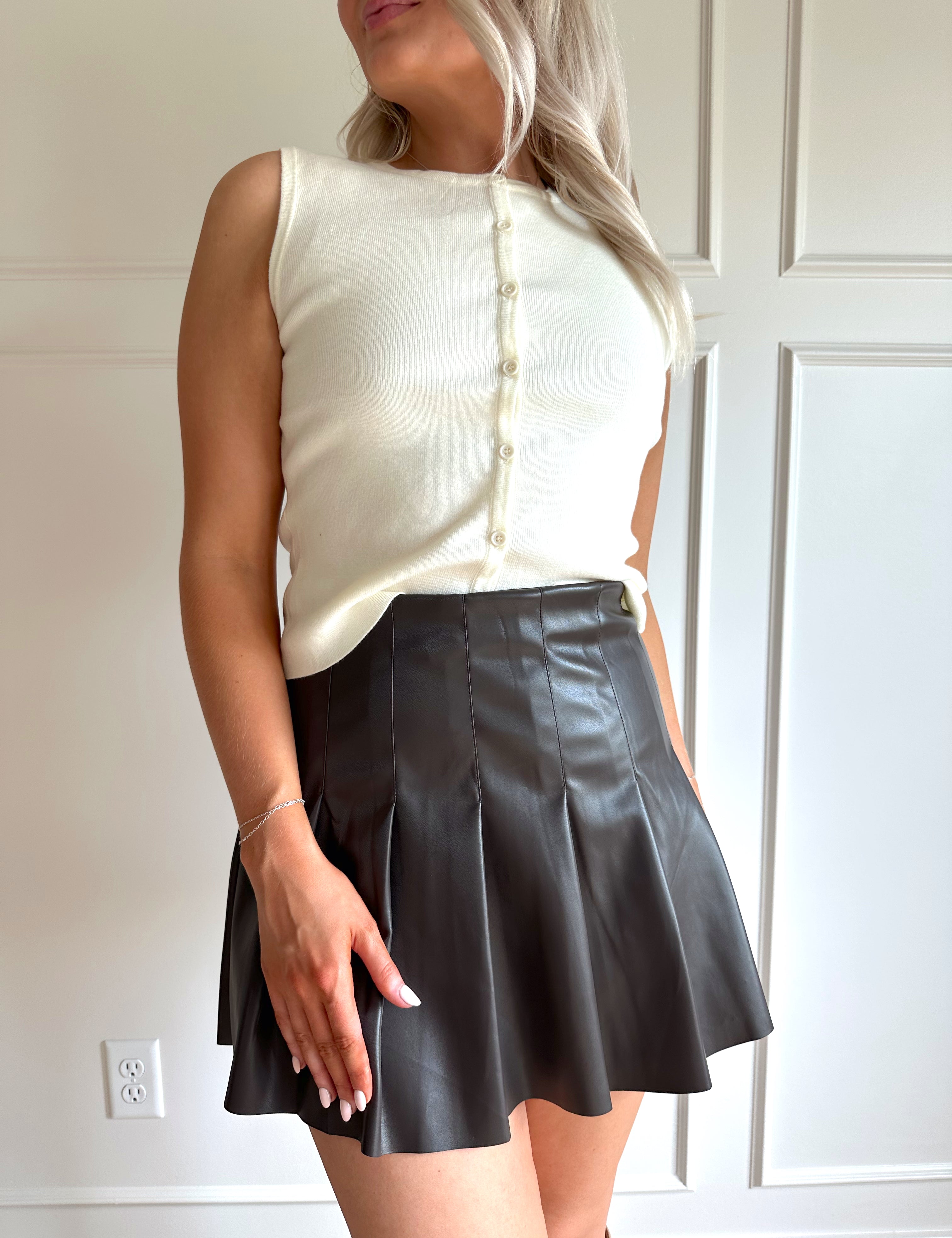 Woman wearing a cream button up sleeveless sweater top with dark brown pleated mini skirt. 