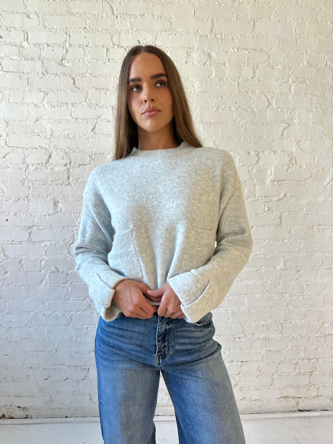 Woman wearing light grey knit sweater with front pocket detail and medium wash wide leg jeans.