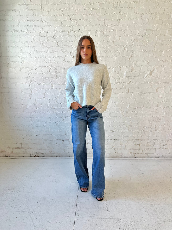 Woman wearing light grey knit sweater with front pocket detail and medium wash wide leg jeans.