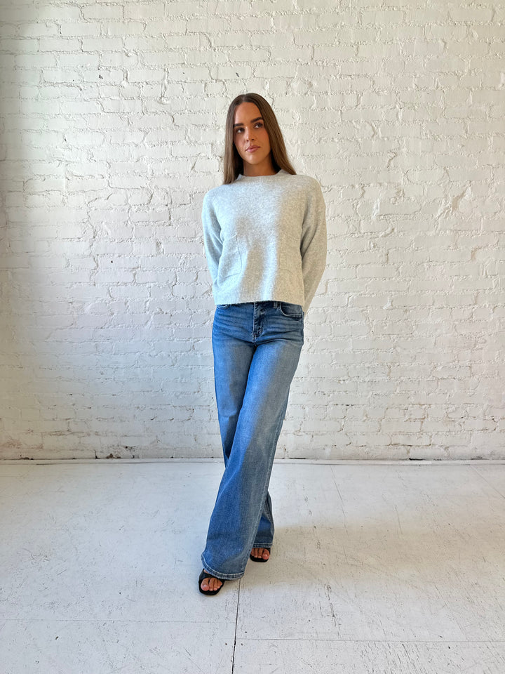 Woman wearing light grey knit sweater with front pocket detail and medium wash wide leg jeans.