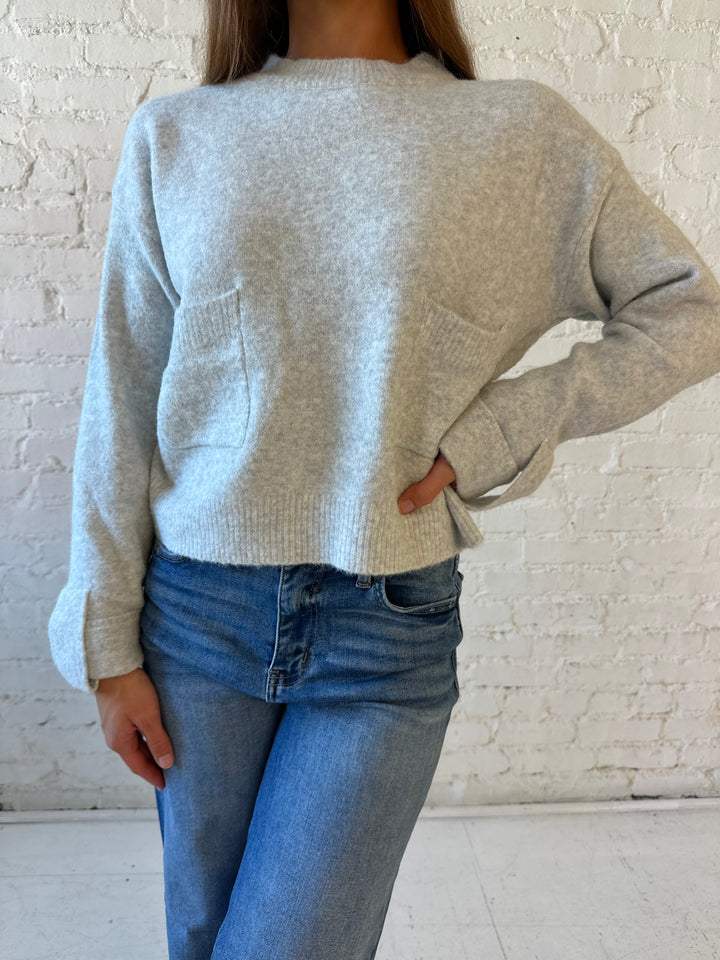Woman wearing light grey knit sweater with front pocket detail and medium wash wide leg jeans.