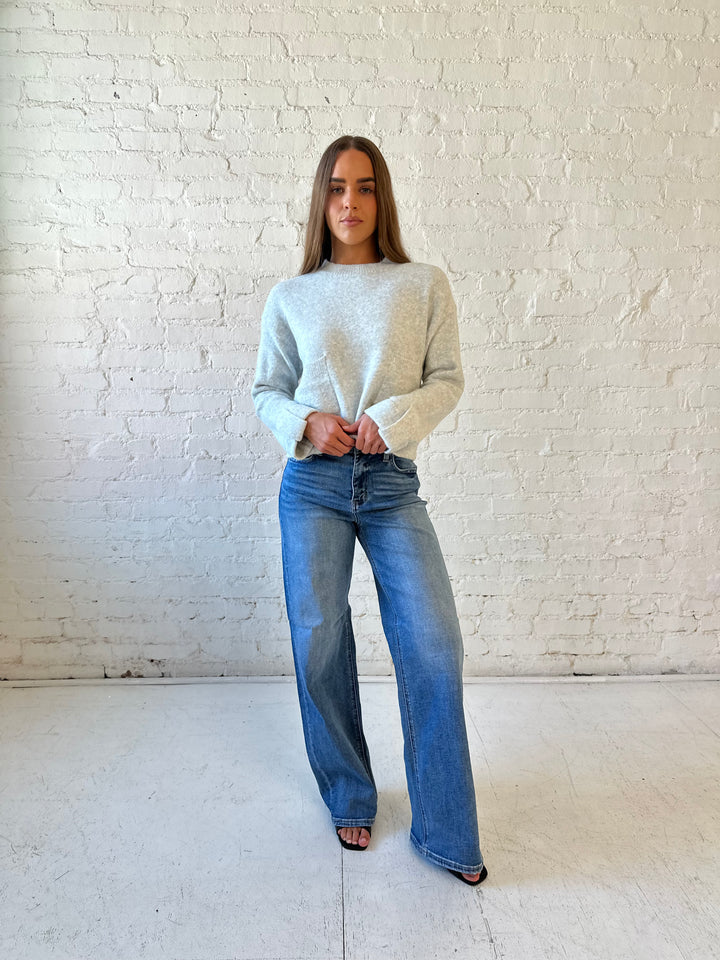 Woman wearing light grey knit sweater with front pocket detail and medium wash wide leg jeans.