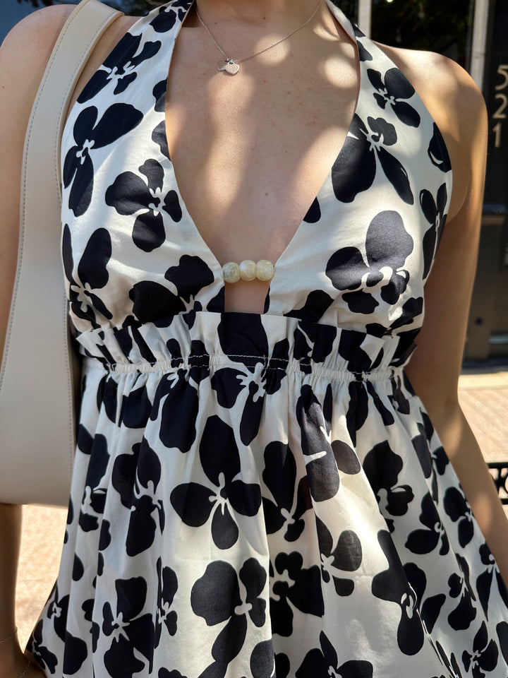 Woman wearing ivory and black floral mini dress with self tie halter neck, beading, and elastic cinched waist. 
