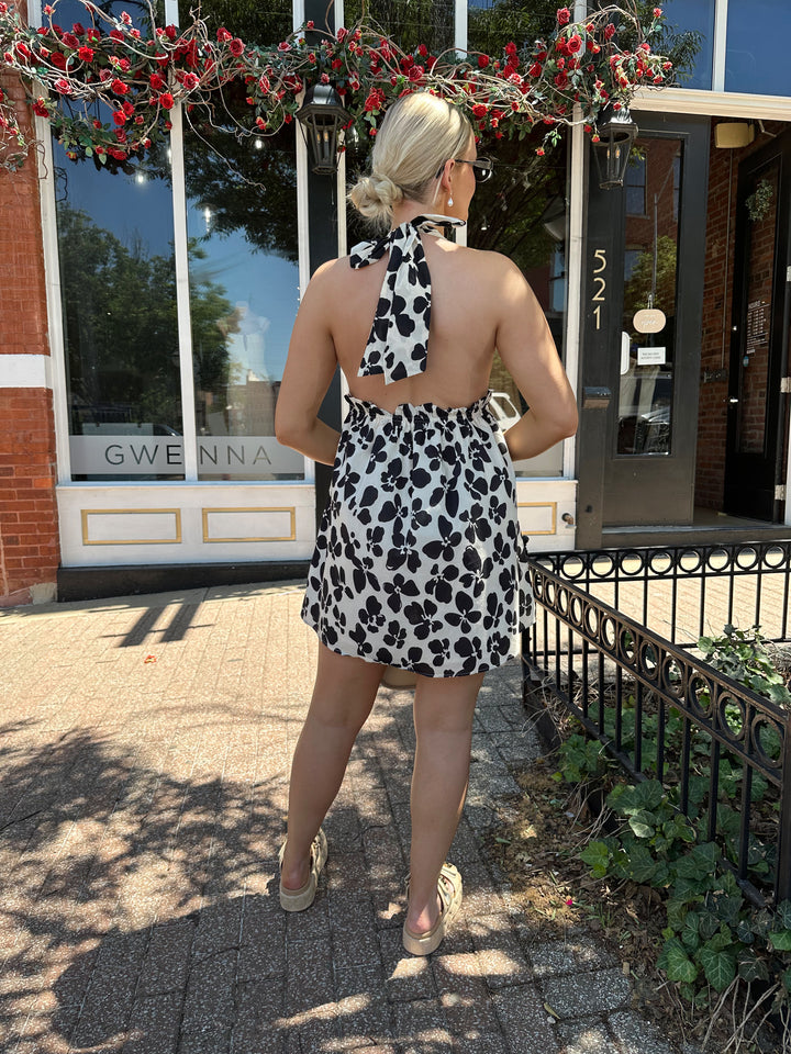 Woman wearing ivory and black floral mini dress with self tie halter neck, beading, and elastic cinched waist. 