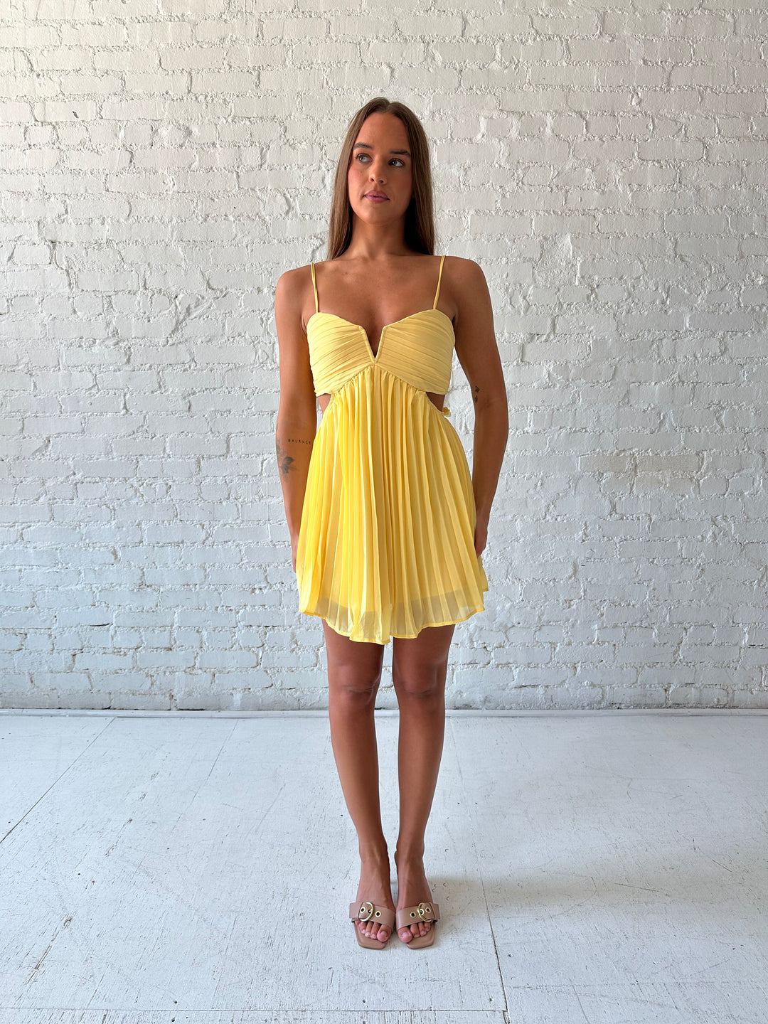 Woman wearing yellow pleated mini dress with side cutouts, and floral tassels. 
