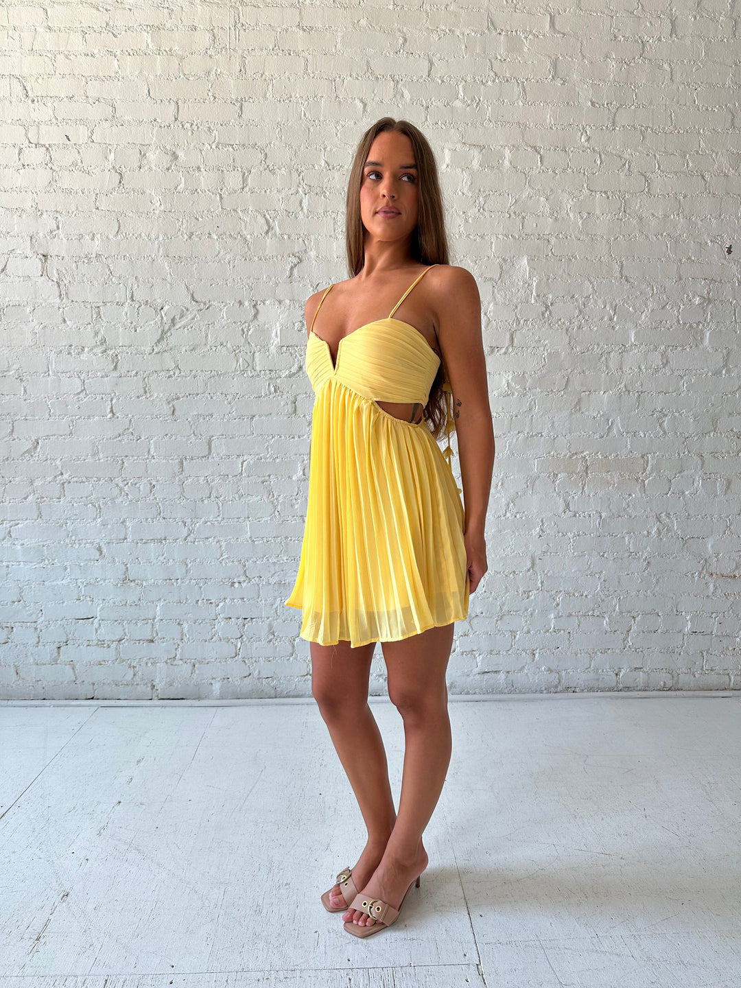 Woman wearing yellow pleated mini dress with side cutouts, and floral tassels. 