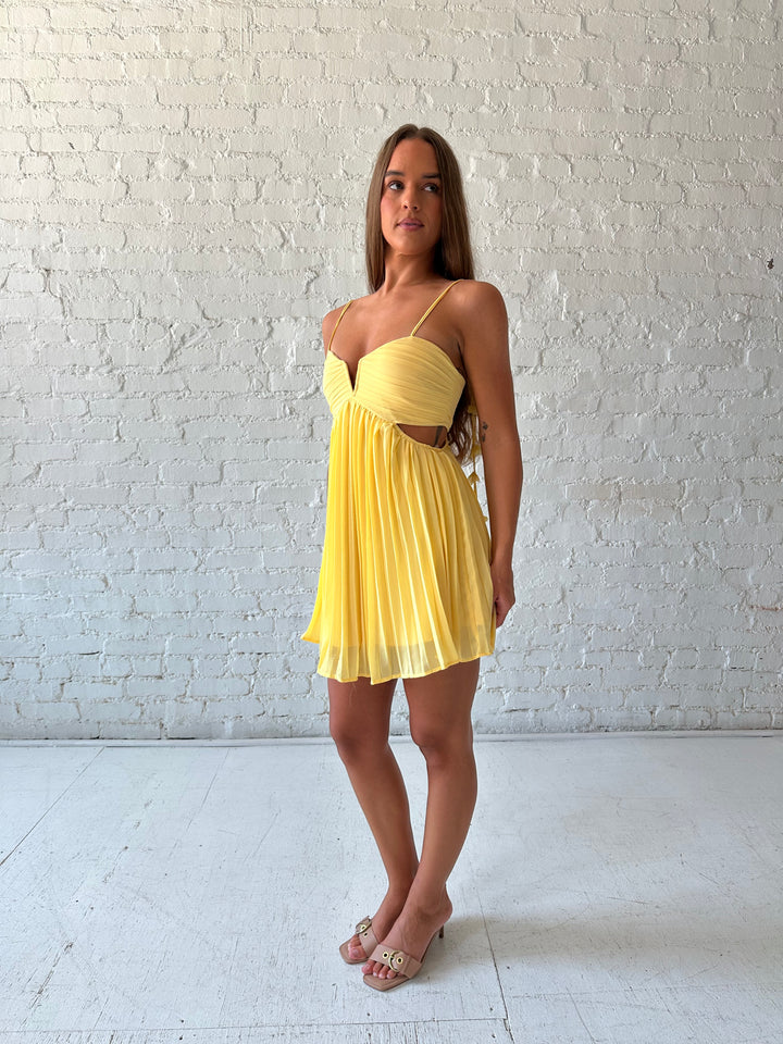 Woman wearing yellow pleated mini dress with side cutouts, and floral tassels. 