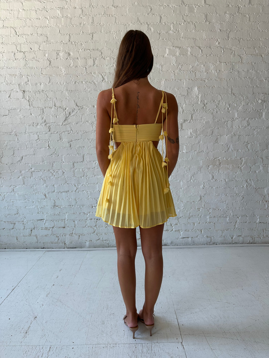 Woman wearing yellow pleated mini dress with side cutouts, and floral tassels. 