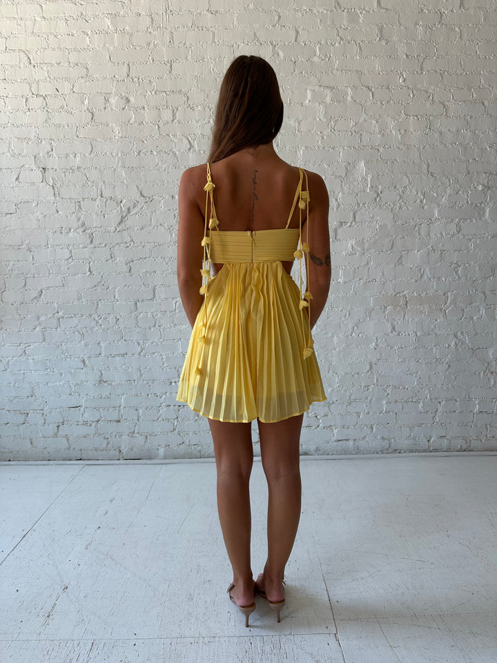 Woman wearing yellow pleated mini dress with side cutouts, and floral tassels. 