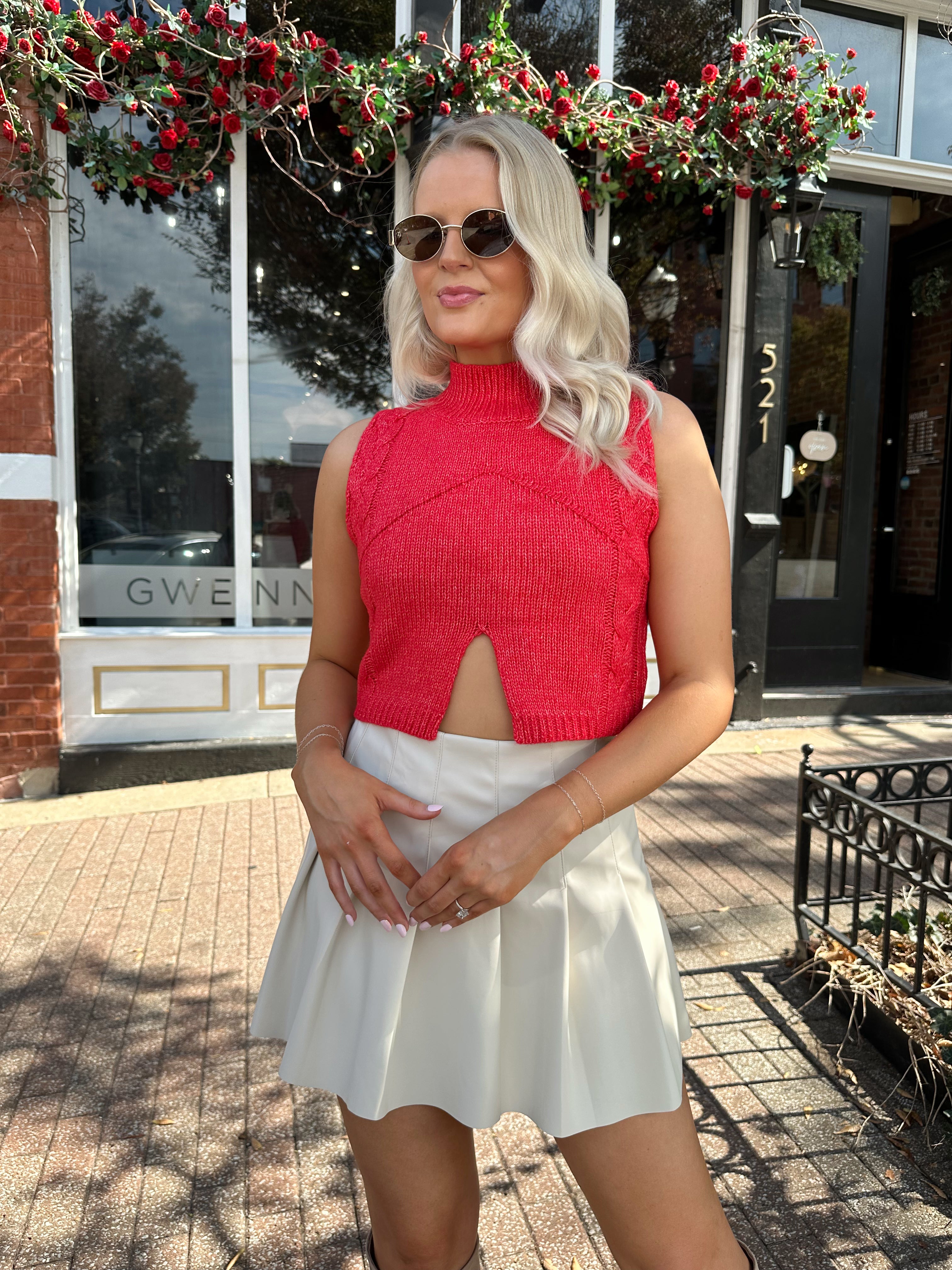 Woman wearing red sleeveless sweater tip with cream pleated leather mini skirt. 