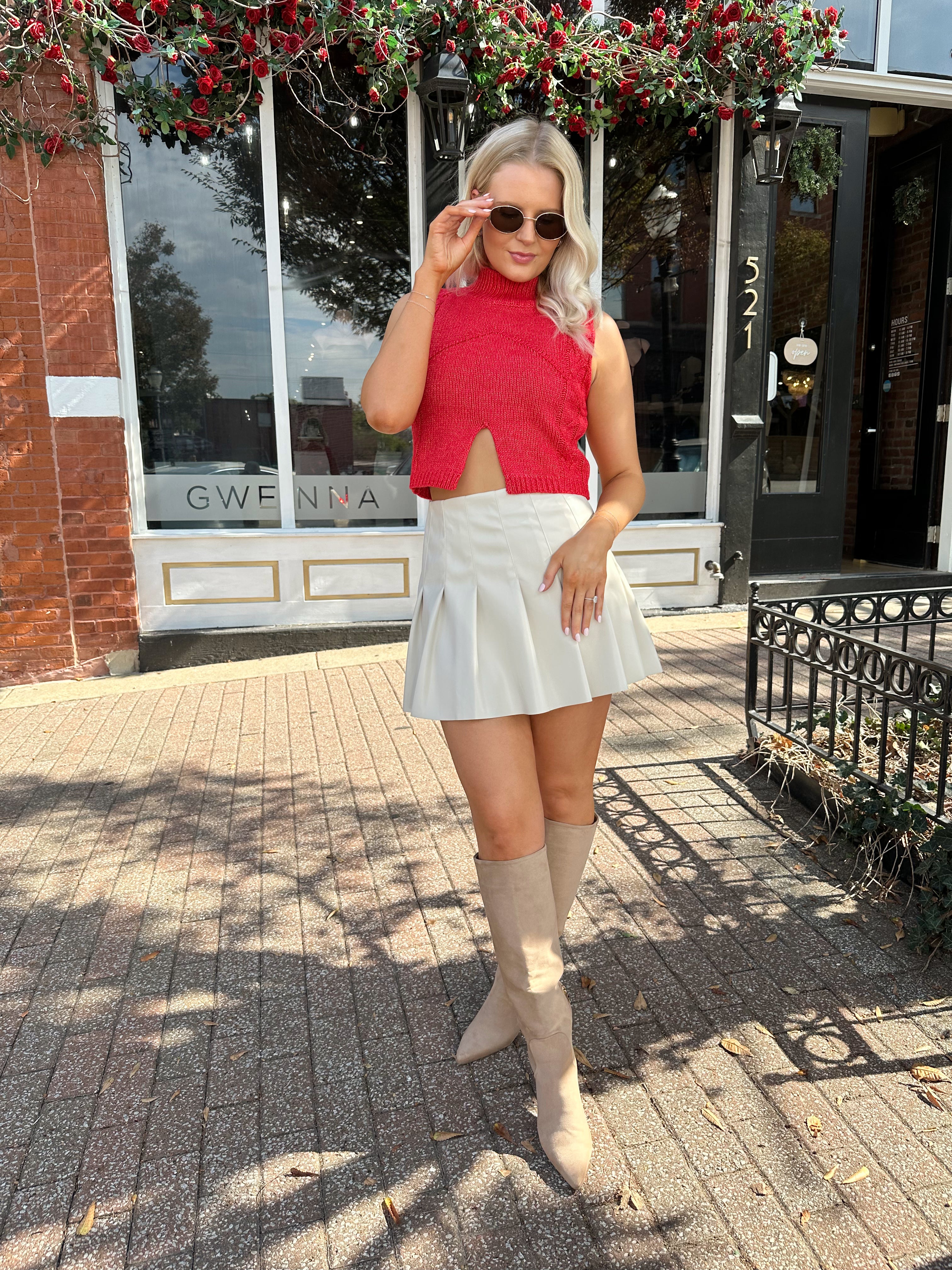 Woman wearing red sleeveless sweater tip with cream pleated leather mini skirt. 