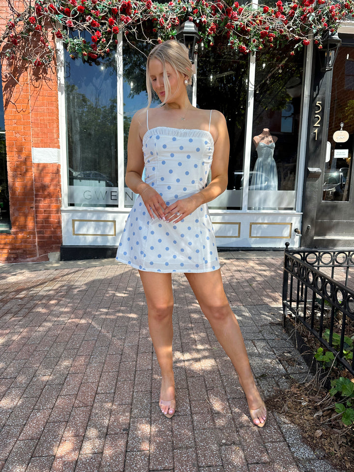 Woman wearing white mini dress with light blue polka dots, and adjustable straps.