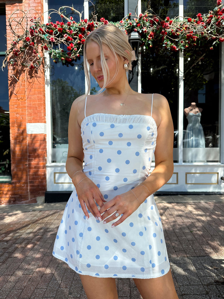 Woman wearing white mini dress with light blue polka dots, and adjustable straps.