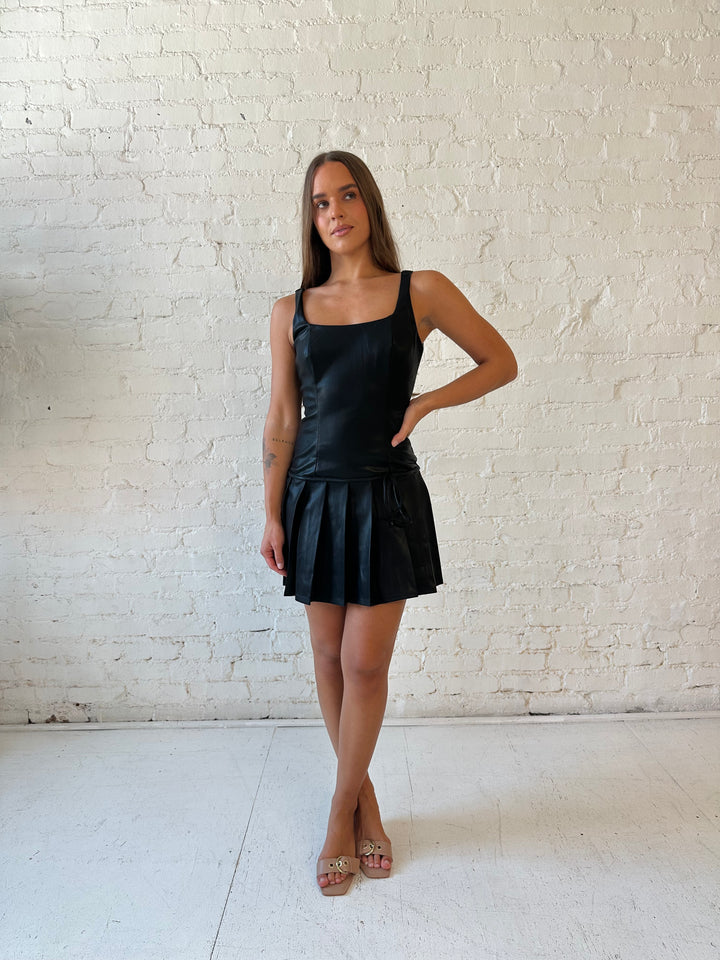 Woman wearing black leather mini dress with pleated bottom and bow tie detail.
