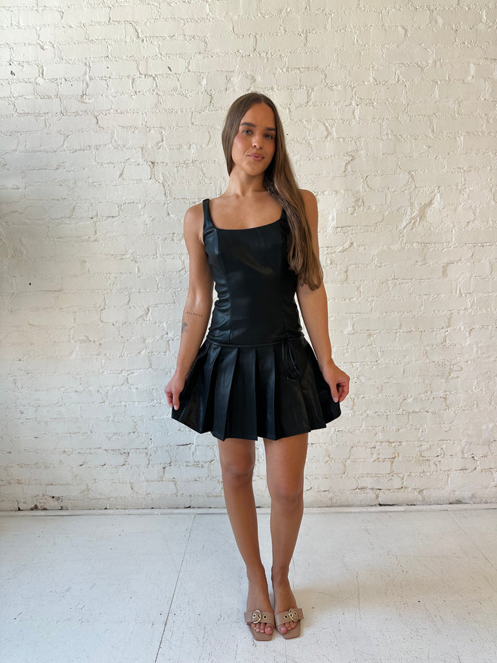 Woman wearing black leather mini dress with pleated bottom and bow tie detail.