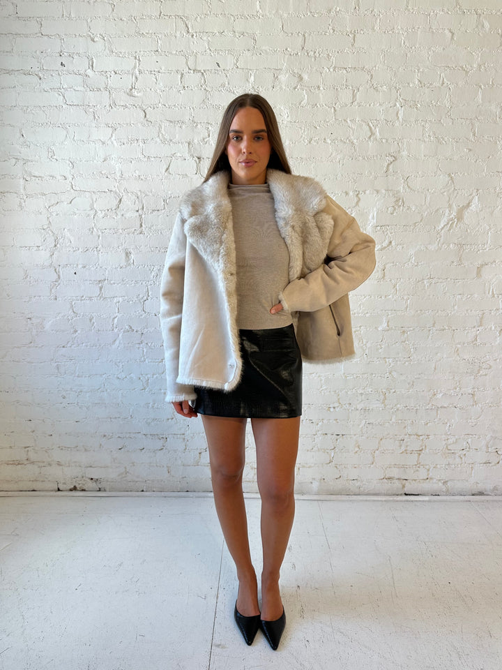 Woman wearing taupe reversible coat with one side being a faux fur, and the other side being suede. 