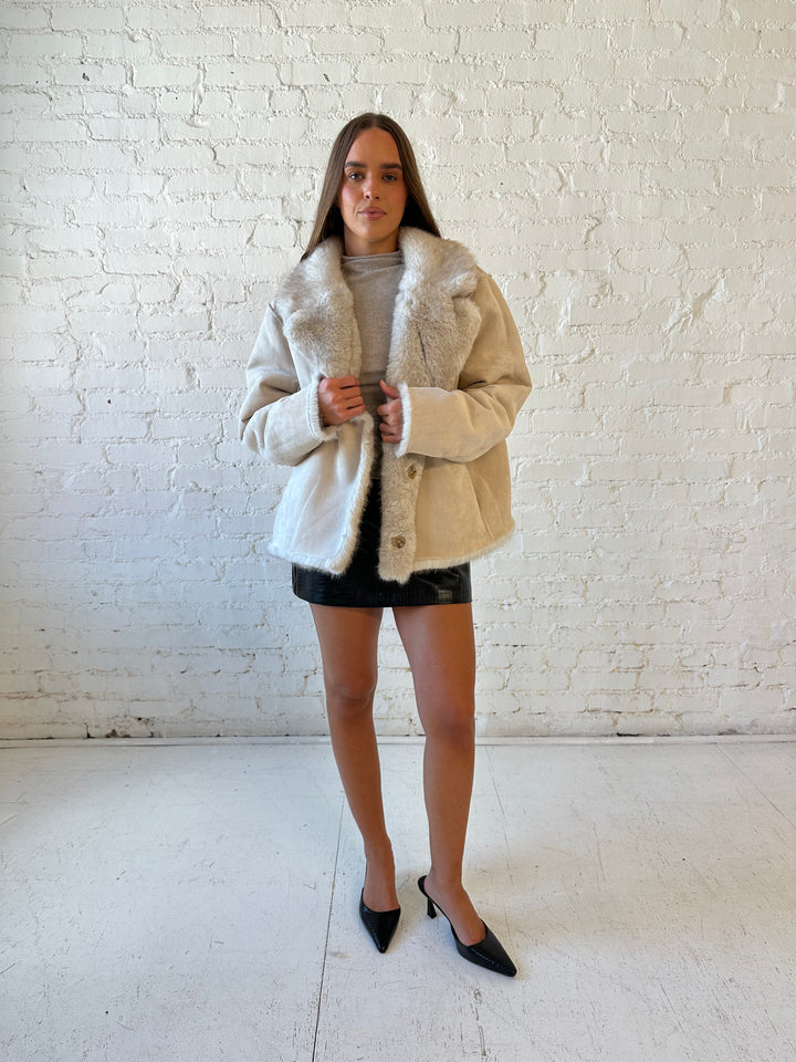 Woman wearing taupe reversible coat with one side being a faux fur, and the other side being suede. 