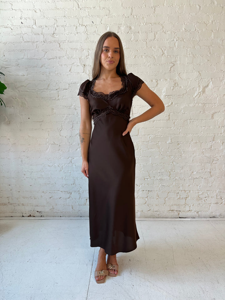 Woman wearing dark brown satin maxi dress with lace trim detailing and flutter sleeves. 
