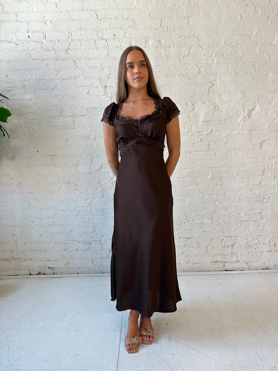 Woman wearing dark brown satin maxi dress with lace trim detailing and flutter sleeves. 