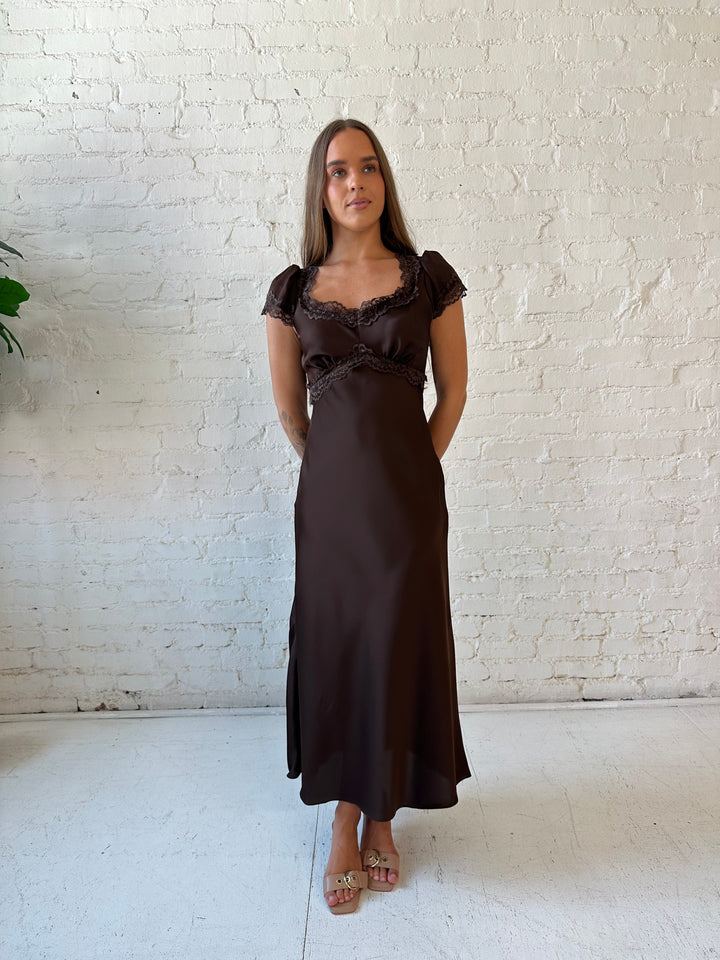 Woman wearing dark brown satin maxi dress with lace trim detailing and flutter sleeves. 