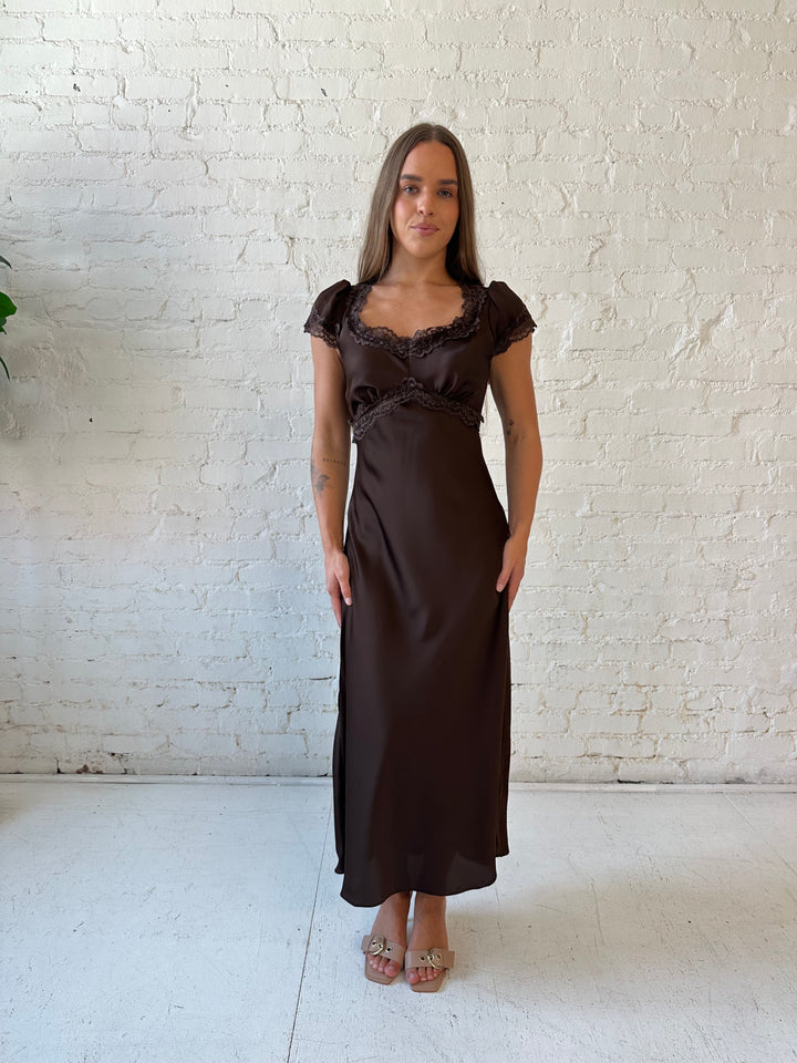 Woman wearing dark brown satin maxi dress with lace trim detailing and flutter sleeves. 