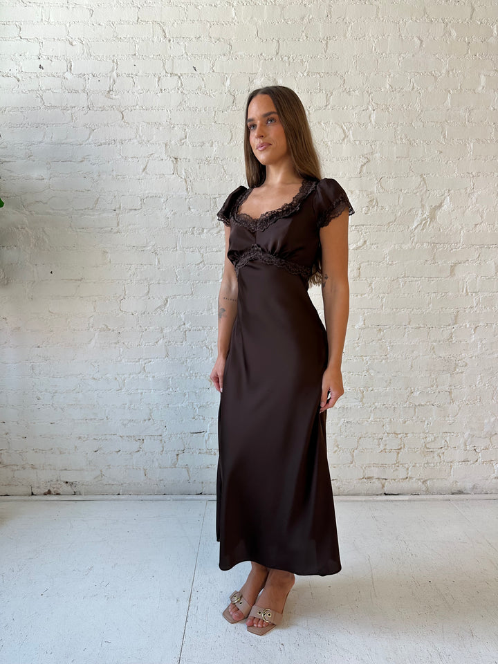 Woman wearing dark brown satin maxi dress with lace trim detailing and flutter sleeves. 