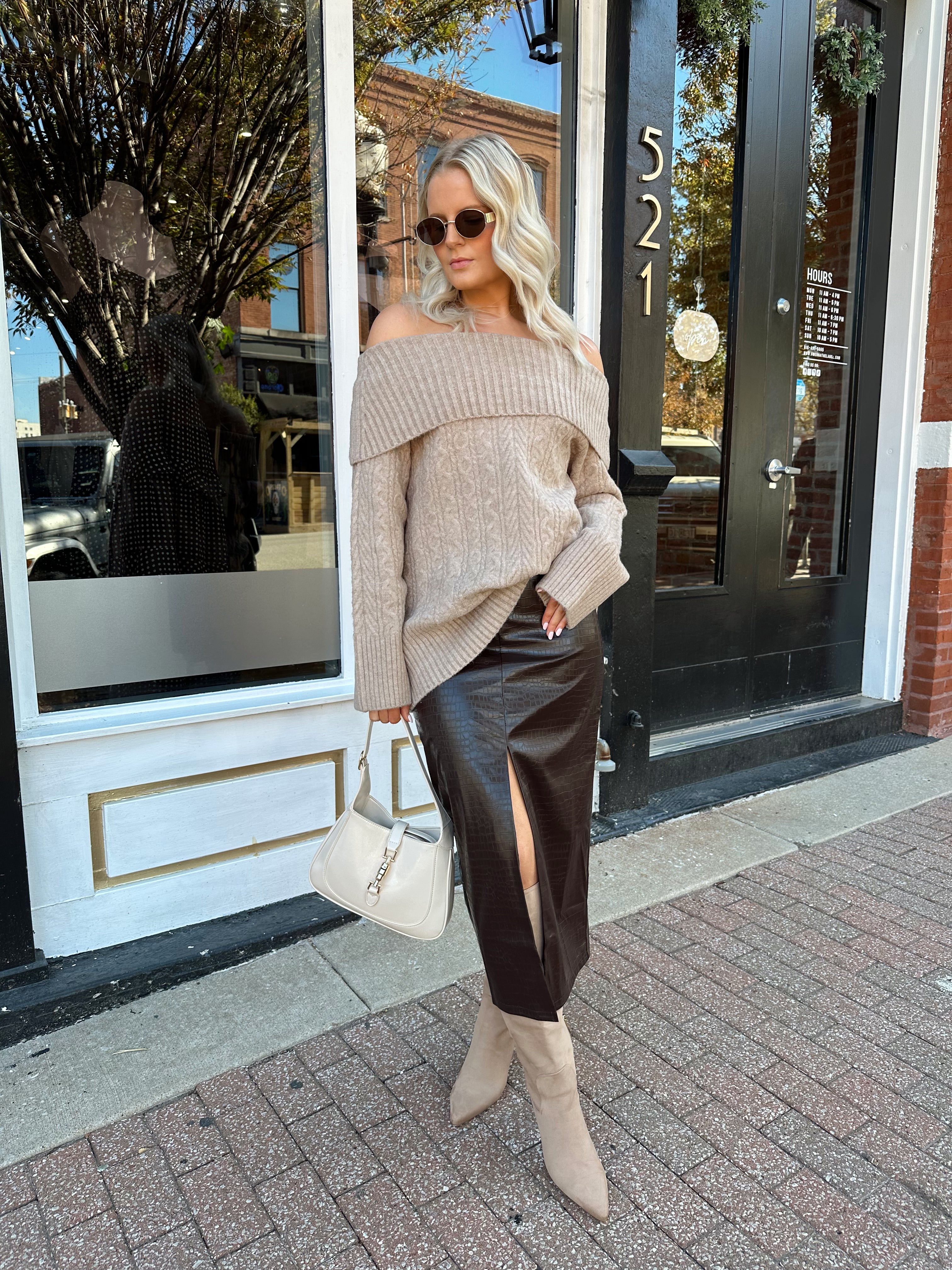 Woman wearing oversized, off the shoulder, tan sweater with brown leather skirt. 