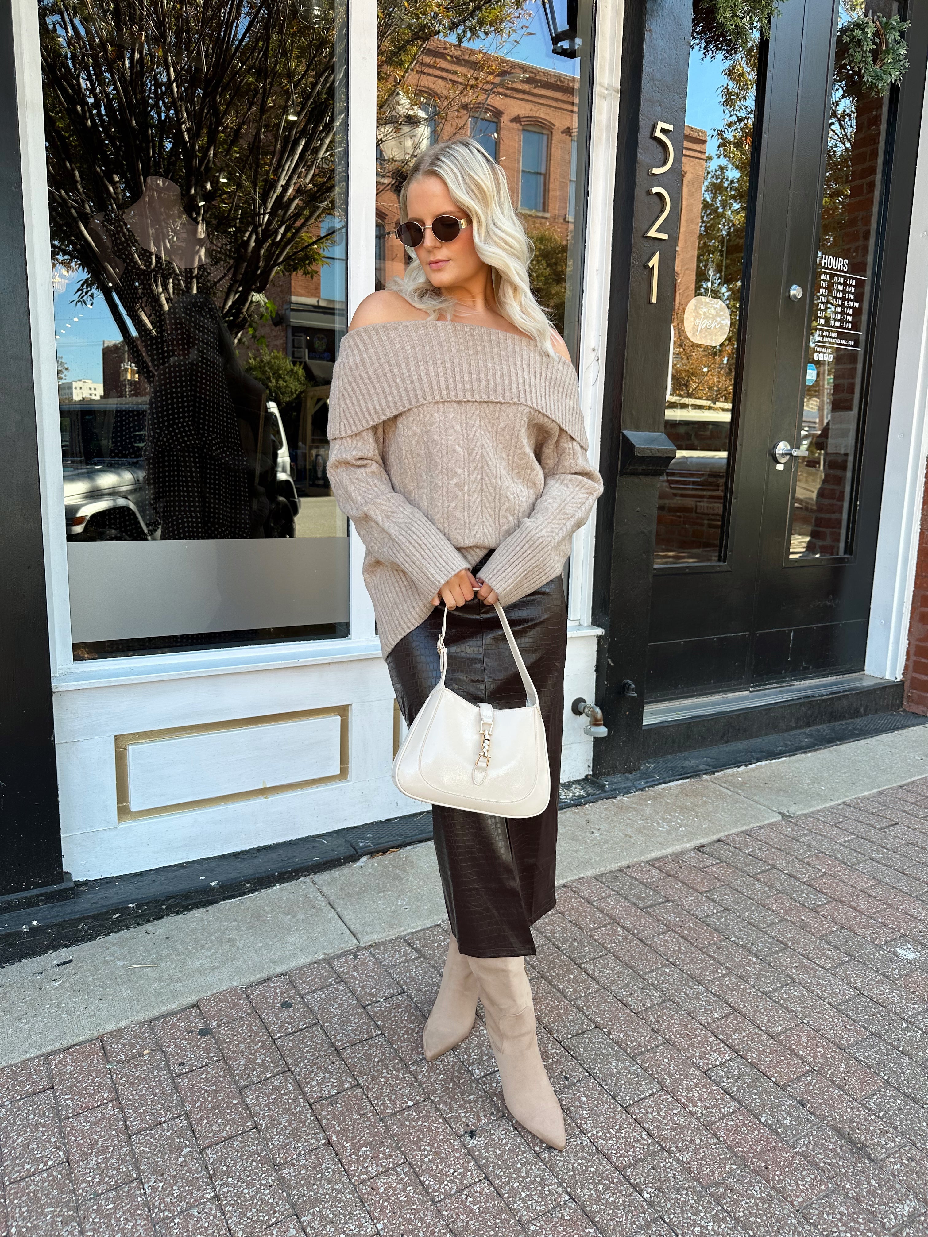 Woman wearing oversized, off the shoulder, tan sweater with brown leather skirt. 