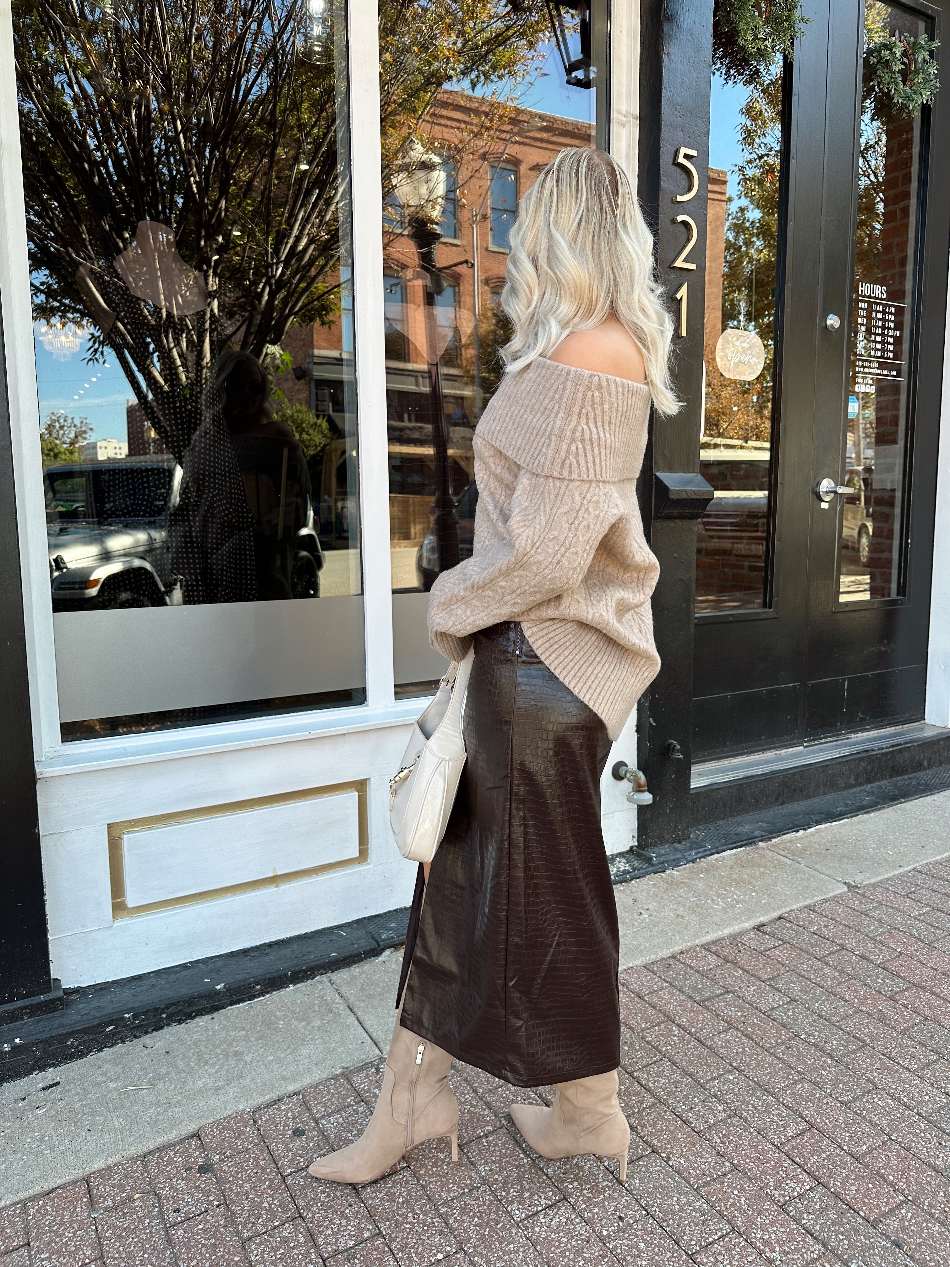 Woman wearing oversized, off the shoulder, tan sweater with brown leather skirt. 