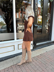 Woman wearing chocolate brown asymmetrical fitted mini dress with tan knee-high boots.