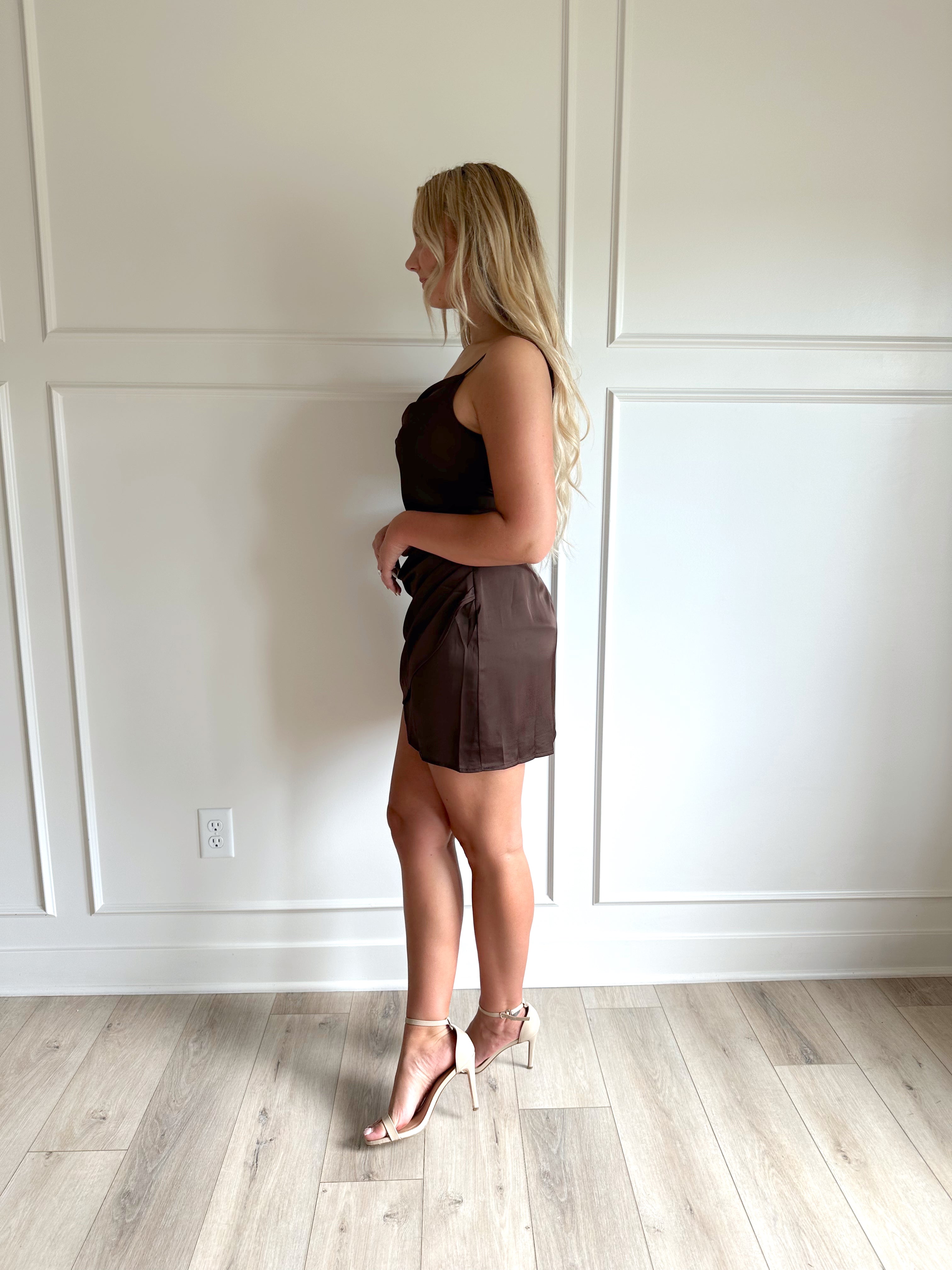 Woman wearing a dark brown satin mini dress with drapery midsection and cowl neckline. 