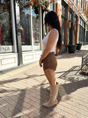 Woman wearing cream knit high neck sleeveless bodysuit and brown suede short. 