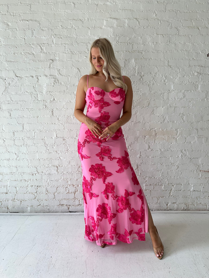 Woman wearing pink maxi dress with floral appliqué detail, and side slit.