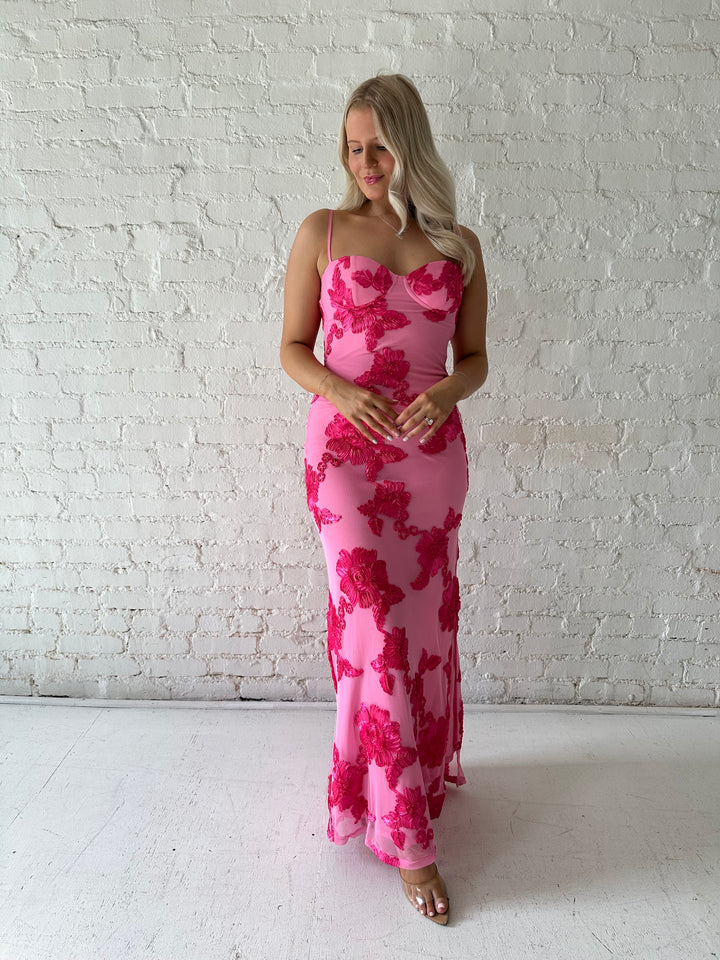 Woman wearing pink maxi dress with floral appliqué detail, and side slit.