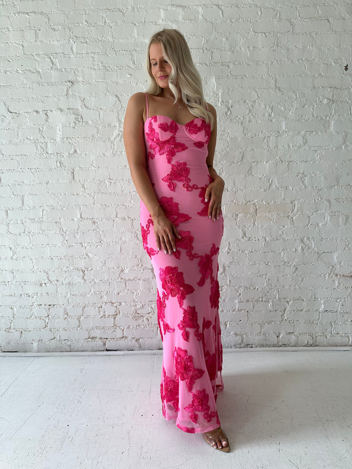 Woman wearing pink maxi dress with floral appliqué detail, and side slit.