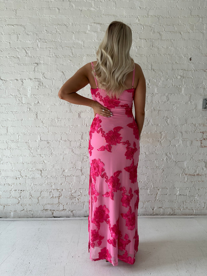 Woman wearing pink maxi dress with floral appliqué detail, and side slit.