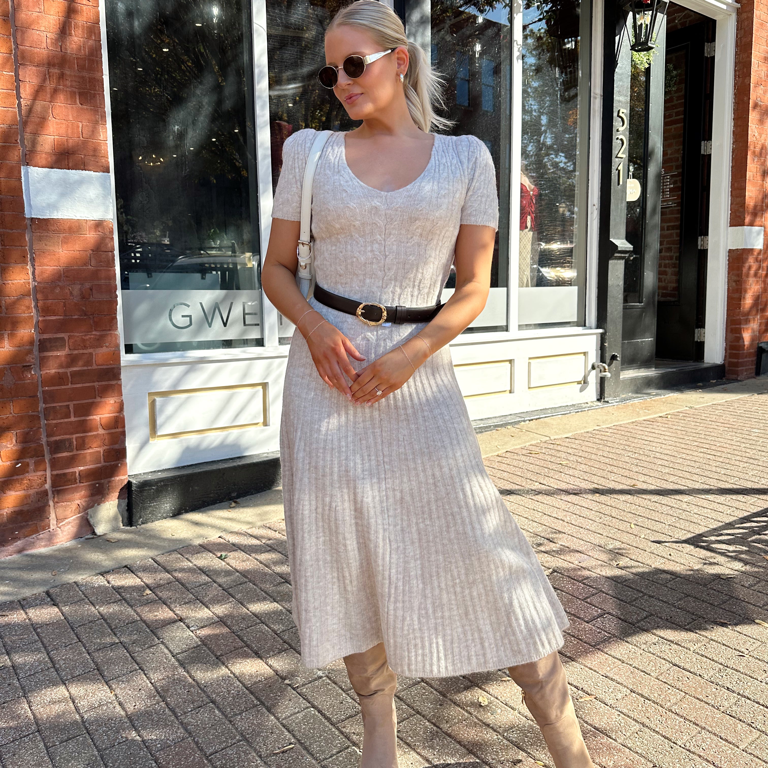 Woman wearing oatmeal cable knit sweater dress with dark belt. 